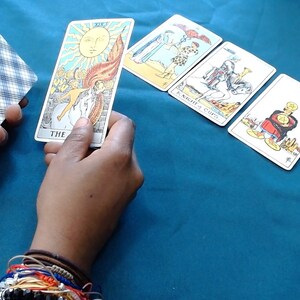 May include: A hand holds a deck of tarot cards with a blue and white chequered pattern. Another hand holds a card depicting the sun. Three other tarot cards are arranged on a teal surface.
