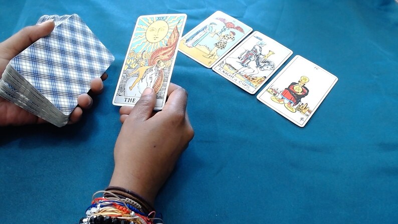 May include: A hand holds a deck of tarot cards with a blue and white chequered pattern. Another hand holds a card depicting the sun. Three other tarot cards are arranged on a teal surface.