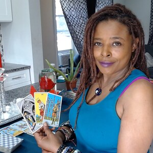 May include: A woman holding tarot cards, with a deck of cards in front of her. The woman is wearing a blue top and jewellery. Red candles in crystal holders are on a silver tray. A glass container with a red flower is visible.