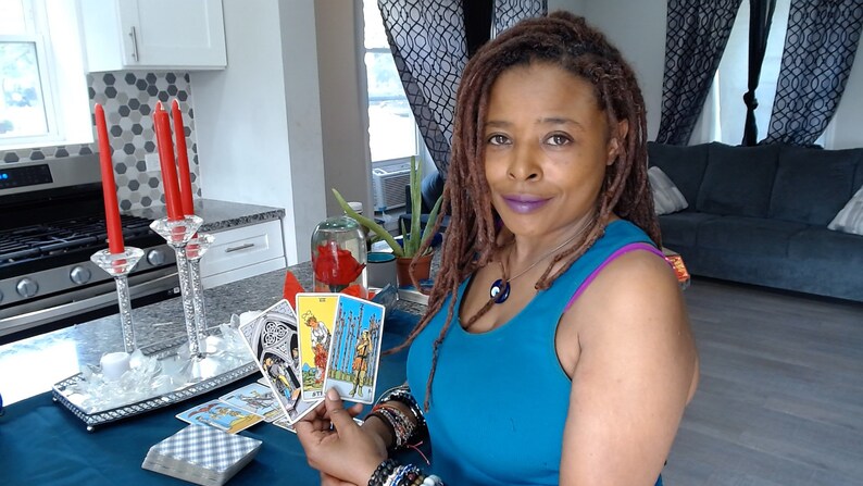 May include: A woman holding tarot cards, with a deck of cards in front of her. The woman is wearing a blue top and jewellery. Red candles in crystal holders are on a silver tray. A glass container with a red flower is visible.