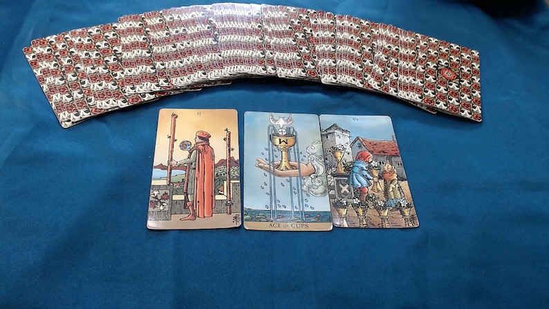 May include: A tarot card spread on a blue surface. The deck's backs have a red and white floral design. Three cards are face up, showing illustrations. The Ace of Cups card is visible, along with other cards from the deck.
