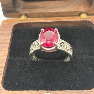 May include: A silver ring featuring a large, oval-cut, deep red gemstone held by four prongs. The band displays an intricate, openwork metal pattern with interconnected loops. The ring is presented inside a dark wooden box with black lining.