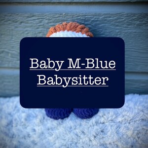 May include: A crocheted plush toy in the shape of a person with brown hair, a white shirt, and blue pants. The toy is sitting on a white fluffy surface. The text "Baby M-Blue Babysitter" is written on a blue rectangle behind the toy.
