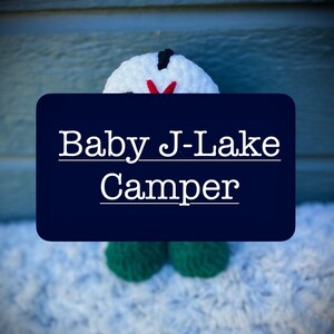 May include: A white crocheted snowman with a red nose and black eyes. The snowman is wearing a green crocheted hat and scarf. The text "Baby J-Lake Camper" is written on a blue background.