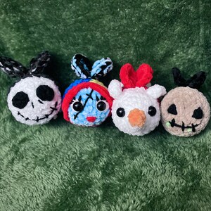 May include: Four crocheted plush toys, each with a different character design. The characters are Jack Skellington, Sally, Oogie Boogie, and a snowman. The toys are all white with different colored accents.
