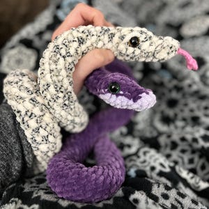 May include: Two crocheted snake plushies. One is white with black speckles and has pink tongue. The other is purple.