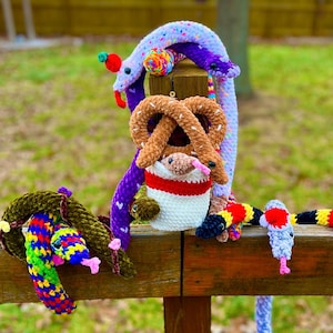 Puede incluir: Una colección de juguetes de serpiente de crochet de colores brillantes, algunos con chispas, están envueltos alrededor de un poste de madera. Un pretzel de crochet marrón y una taza de crochet con un diseño de rayas rojas y blancas también están en el poste.