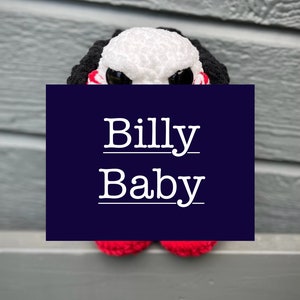 May include: Crocheted plush toy of Billy the puppet from the Saw horror movie franchise. The toy is white with black hair and red accents. The toy has large black eyes and a red smile. The toy is sitting on a gray surface. The text "Billy Baby" is written in white on a blue background.