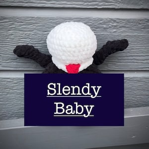 May include: A crocheted Slendy Baby plush toy with a white head, black arms and legs, and a red mouth. The toy is sitting on a gray wooden surface with a blue sign that says "Slendy Baby".
