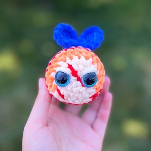May include: A crocheted toy with a red, orange, and white body and blue bow. The toy has large blue eyes.