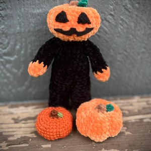 May include: A crocheted pumpkin character with a black body and orange pumpkin head. The character is standing on a wooden surface with two smaller orange pumpkins.