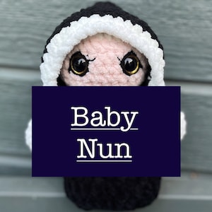 Puede incluir: Una muñeca de bebé de crochet que lleva una túnica negra con capucha y ribete blanco. La muñeca tiene grandes ojos negros y una cara rosa. La muñeca sostiene un cartel que dice "Baby Nun".