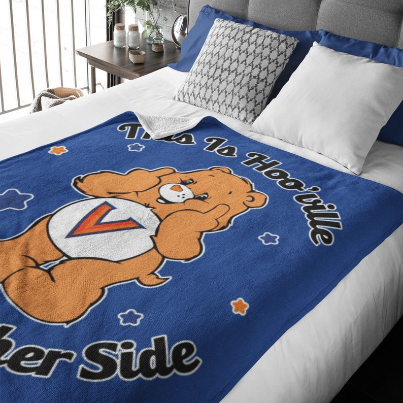 Puede incluir: Una manta de forro polar azul con un oso de dibujos animados que lleva una camiseta con una "V" en ella. El texto "This is Hooville" est&aacute; escrito en un arco curvo sobre el oso. El texto "er Side" est&aacute; escrito debajo del oso.