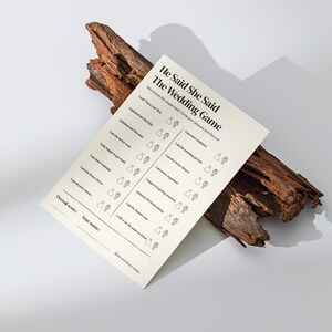 Könnte beinhalten: Eine cremefarbene Hochzeitsspielkarte mit dem Titel "He Said She Said The Wedding Game" liegt auf einem Stück dunkelbraunem Holz. Die Karte enthält eine Liste von Fragen und Platz für Antworten. Der Hintergrund ist eine schlichte weiße Oberfläche.