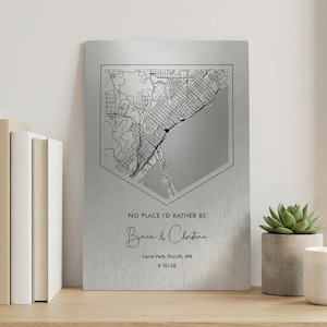 Könnte beinhalten: Silberfarbener Metall-Karten-Druck mit einem schwarz-weißen Kartendesign in einer geometrischen Form. Der Text lautet "NO PLACE I'D RATHER BE", mit den Namen "Brian & Christian" und dem Datum "6-30-18".