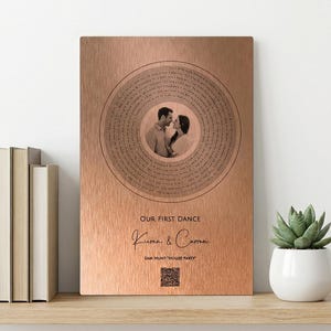 Pode incluir: Placa de metal cor de cobre com uma foto circular de um casal, rodeada por texto. A placa diz "Our First Dance" com nomes abaixo e um código QR. Uma pequena planta suculenta num vaso branco está numa prateleira de madeira.