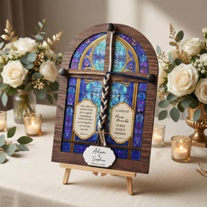 May include: A wooden framed stained glass art piece with a blue and purple design. The art piece has a braided rope and a small tag with the names "Adam & Sophia" and the date "February 12, 2026". The art is displayed on a small wooden easel.