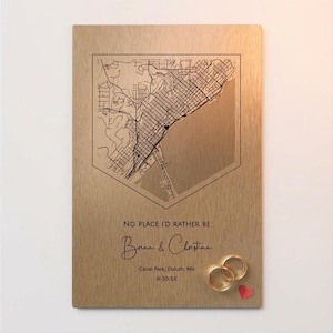 May include: A gold-colored metal plaque with a detailed map of a city, framed within a geometric shape. The text "NO PLACE I'D RATHER BE" is above the names "Brian & Christian". Two gold rings and a red heart are at the bottom.