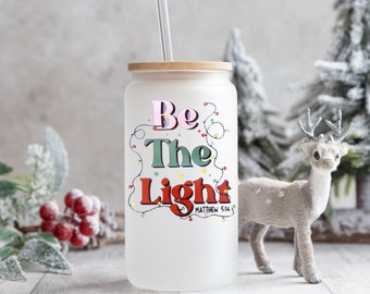 May include: A frosted glass tumbler with a bamboo lid and a clear straw. The tumbler features the text "Be The Light" in colorful lettering with string lights. A decorative reindeer figurine is in the background.