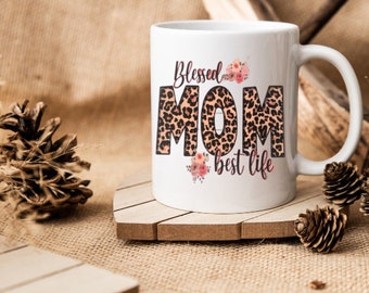 May include: White ceramic mug with the words "Blessed MOM best life" in a mix of fonts and colors. The word "MOM" has a leopard print design. Floral accents are placed around the text. The mug is on a wooden coaster.