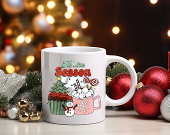 May include: White ceramic mug with a festive design. The mug features the text "Tis the Season" with illustrations of a Christmas tree, cupcake, snowman, and a mug of hot cocoa. Red and green accents.