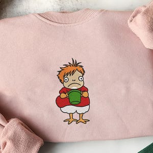 May include: A light pink sweatshirt featuring an embroidered cartoon character. The character has orange hair, a red shirt, white shorts, and yellow chicken feet, holding a green bucket. The sweatshirt has a crew neck and long sleeves.