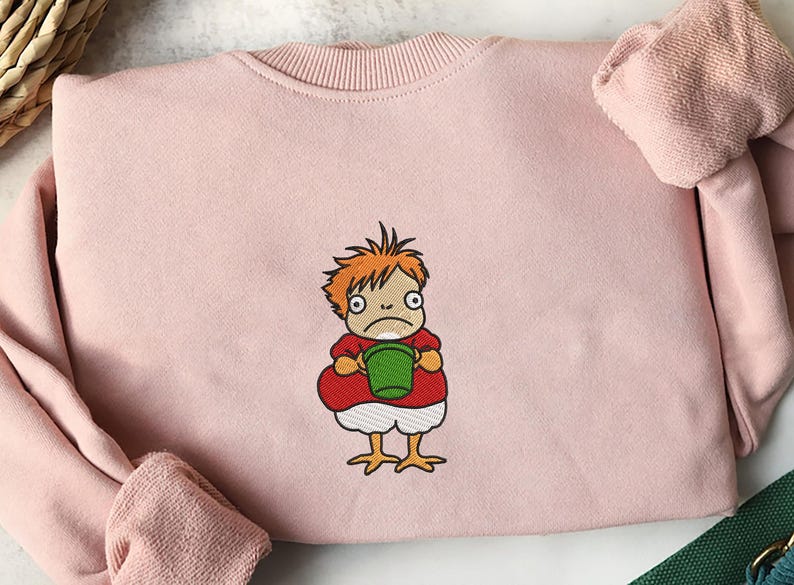 May include: A light pink sweatshirt featuring an embroidered cartoon character. The character has orange hair, a red shirt, white shorts, and yellow chicken feet, holding a green bucket. The sweatshirt has a crew neck and long sleeves.