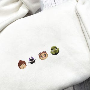 May include: Cream-colored sweatshirt featuring embroidered cartoon character faces in brown, black, tan, and green. The embroidered designs are arranged in a horizontal line across the chest area of the sweatshirt.