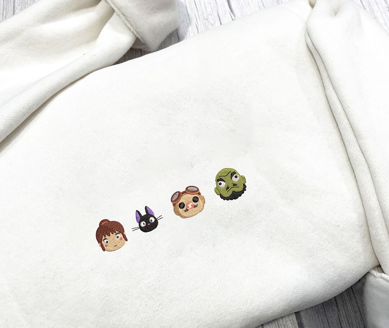 May include: Cream-colored sweatshirt featuring embroidered cartoon character faces in brown, black, tan, and green. The embroidered designs are arranged in a horizontal line across the chest area of the sweatshirt.
