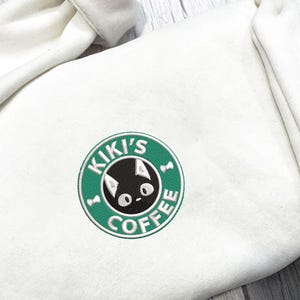 K&ouml;nnte beinhalten: Cremefarbener Kapuzenpullover mit einem gestickten Aufn&auml;her. Der Aufn&auml;her ist ein gr&uuml;ner Kreis mit den Worten "KIKI'S COFFEE" und einem schwarzen Katzenkopf in der Mitte. Der Kapuzenpullover hat eine Kapuze und eine weiche, strukturierte Optik.