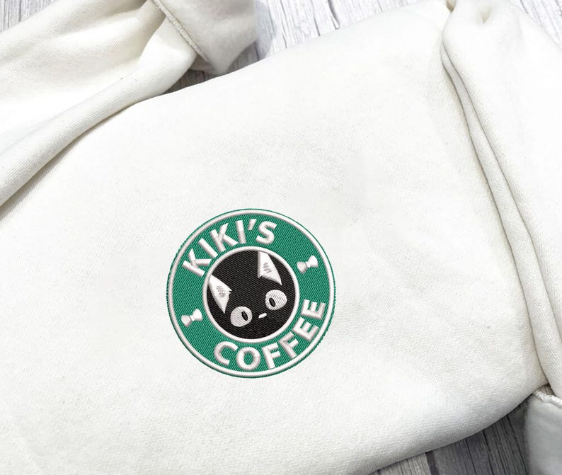 K&ouml;nnte beinhalten: Cremefarbener Kapuzenpullover mit einem gestickten Aufn&auml;her. Der Aufn&auml;her ist ein gr&uuml;ner Kreis mit den Worten "KIKI'S COFFEE" und einem schwarzen Katzenkopf in der Mitte. Der Kapuzenpullover hat eine Kapuze und eine weiche, strukturierte Optik.