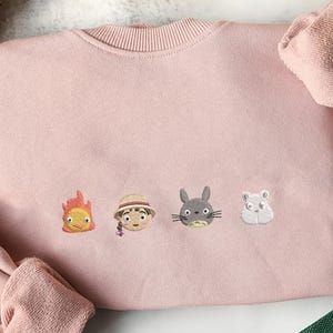 May include: A light pink sweatshirt with embroidered cartoon character faces. The characters include a flame, a girl in a hat, a gray rabbit, and a white hamster. The sweatshirt has a ribbed crew neck and cuffs.