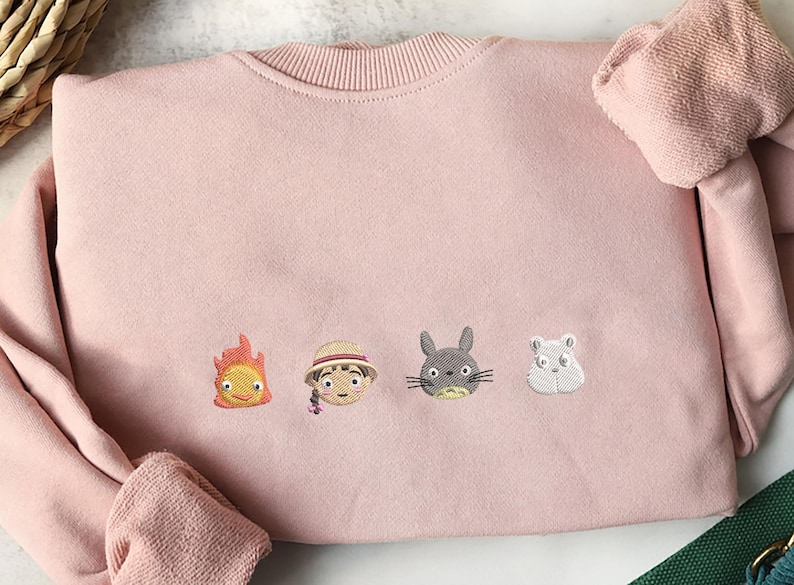 May include: A light pink sweatshirt with embroidered cartoon character faces. The characters include a flame, a girl in a hat, a gray rabbit, and a white hamster. The sweatshirt has a ribbed crew neck and cuffs.
