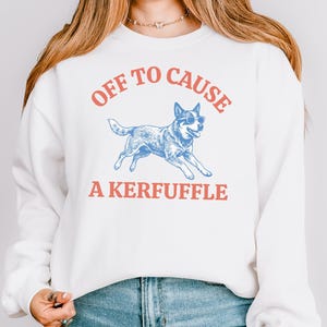 May include: White sweatshirt with the text "OFF TO CAUSE A KERFUFFLE" in red and blue. A blue dog wearing sunglasses is in the center of the design. The sweatshirt has long sleeves and a crew neck.
