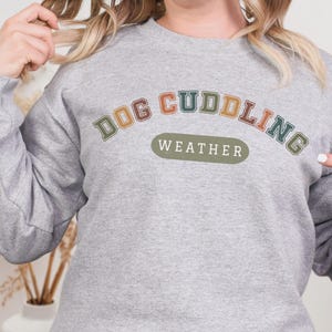 May include: A light gray sweatshirt with the text "DOG CUDDLING WEATHER" in a colorful, collegiate-style font. The sweatshirt is a long-sleeved, crew neck style.