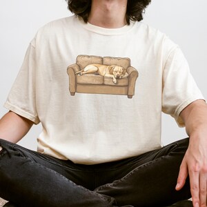 May include: Cream-colored t-shirt featuring a graphic of a light brown dog sleeping on a brown couch. The dog is lying down with its head resting on the arm of the couch. The t-shirt is a relaxed fit.