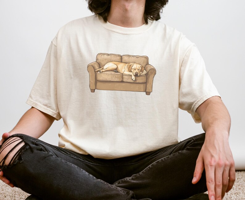 May include: Cream-colored t-shirt featuring a graphic of a light brown dog sleeping on a brown couch. The dog is lying down with its head resting on the arm of the couch. The t-shirt is a relaxed fit.