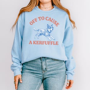 May include: Light blue sweatshirt with the text "OFF TO CAUSE A KERFUFFLE" in red. A blue and white dog wearing sunglasses is in the center. The sweatshirt is paired with blue jeans.