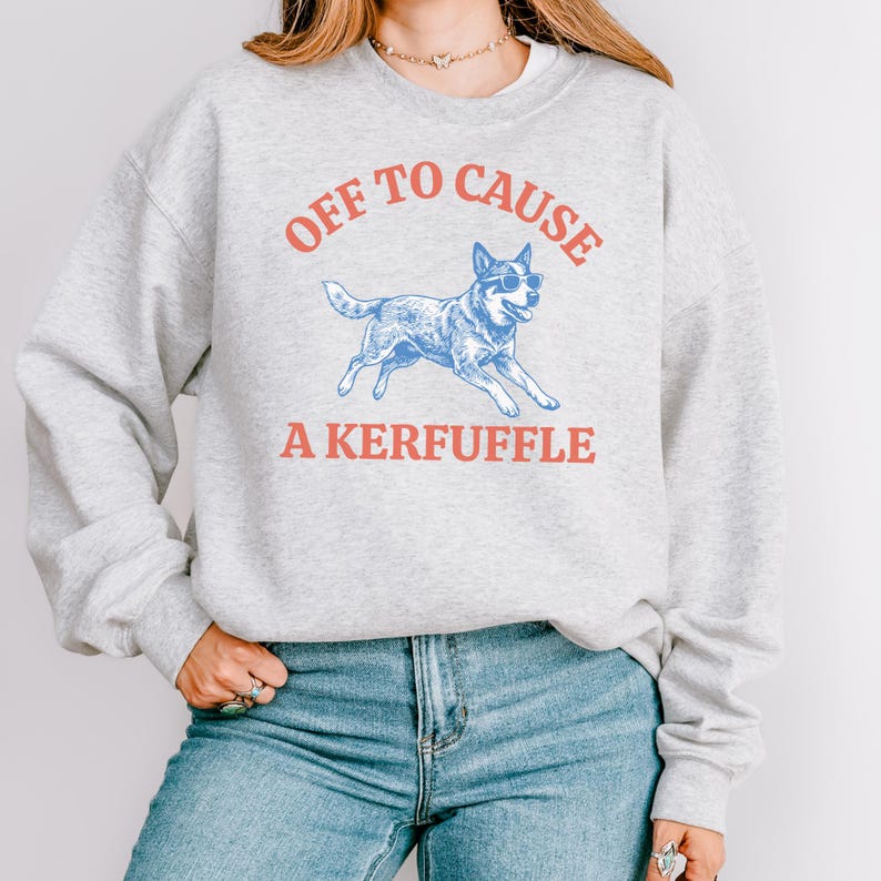 May include: A light gray sweatshirt with the text "OFF TO CAUSE A KERFUFFLE" in red and a blue illustration of a dog wearing sunglasses. The sweatshirt is a casual style.