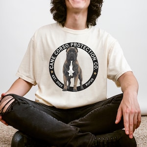 May include: Cream-colored t-shirt featuring a circular graphic with a Cane Corso dog illustration. The graphic includes the text "Cane Corso Protection Co." and "Est. 2025 Rufus". The dog is brown and black with a white chest.