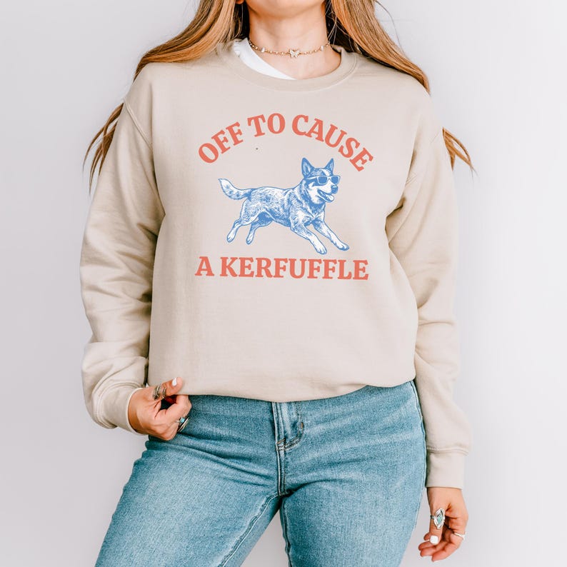 May include: Beige sweatshirt with the text "OFF TO CAUSE A KERFUFFLE" in red and blue. A blue dog wearing sunglasses is in the center. The sweatshirt is long-sleeved and has a crew neck.