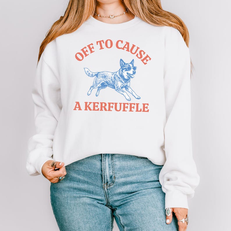 May include: White sweatshirt with the text "OFF TO CAUSE A KERFUFFLE" in red and blue. A blue dog wearing sunglasses is in the center of the design. The sweatshirt has long sleeves and a crew neck.