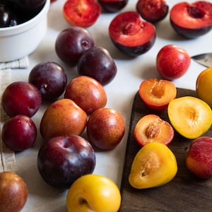 May include: A collection of fresh plums in various colors, including red, purple, and yellow. Some plums are whole, while others are halved, revealing their juicy interiors. A white bowl holds more plums, and a knife rests nearby.