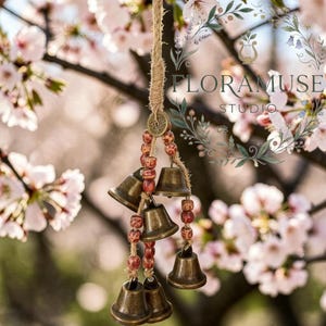 May include: A decorative wind chime featuring small bronze bells and red and brown beads, suspended from a natural rope. The bells are clustered together, and the background shows soft pink blossoms and the text "FLORAMUSE STUDIO."