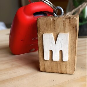 May include: A wooden keychain featuring the letter "M" painted in white. The keychain is attached to a red car key.