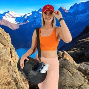 May include: A woman wearing a red baseball cap with a camel logo, an orange sports bra, and pink leggings, is holding a black duffel bag with two water bottles inside. The bag has a black strap and a zipper closure. The woman is standing in front of a mountain range.