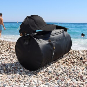 May include: Black duffel bag with a black baseball cap on top. The bag has a gold zipper pull and the text "GYMKILANEC" printed on the side. The bag is sitting on a beach of small pebbles.
