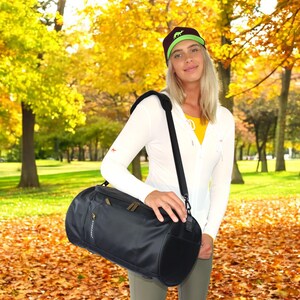 May include: A black cylindrical duffel bag with a shoulder strap. The bag has a zippered pocket on the front and a gold zipper pull. The bag is being held by a person wearing a white sweater and a green and black baseball cap.