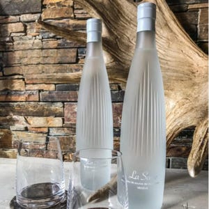 May include: Two frosted glass water bottles with white caps and vertical etched lines, alongside three clear drinking glasses. The bottles have the text "La Source" and "MEGEVE" printed on them. The background features a stone wall and a piece of driftwood.