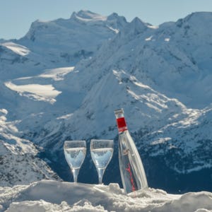 Puede incluir: Una botella de vidrio transparente con etiqueta roja y tapón plateado junto a dos copas de tallo largo llenas de líquido transparente. Los objetos están colocados sobre nieve blanca frente a picos montañosos nevados bajo un cielo azul.